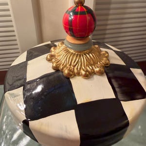 May include: A ceramic jar lid featuring a black and white checkered pattern. The lid is topped with a gold-colored decorative base and a spherical finial painted with a red, green, and black plaid design, finished with a small gold-colored ball.
