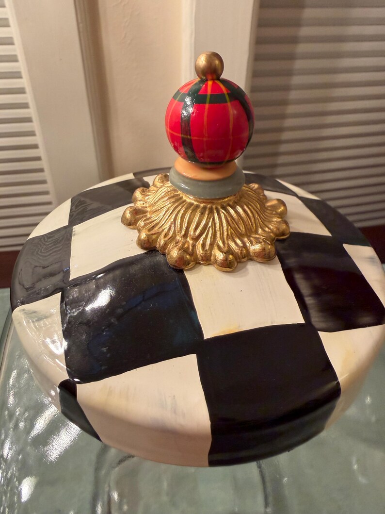 May include: A ceramic jar lid featuring a black and white checkered pattern. The lid is topped with a gold-colored decorative base and a spherical finial painted with a red, green, and black plaid design, finished with a small gold-colored ball.