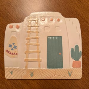 May include: Southwestern-style ceramic wall plaque in the shape of a pueblo. The off-white building features a teal door, a ladder, and a cactus. Decorative elements include a hanging textile and a textured base.