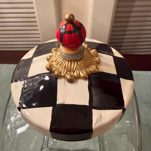 May include: A glass jar lid featuring a black and white checkered pattern. The lid is topped with a gold-colored decorative base and a small, spherical finial painted with a red, green, and black plaid design.
