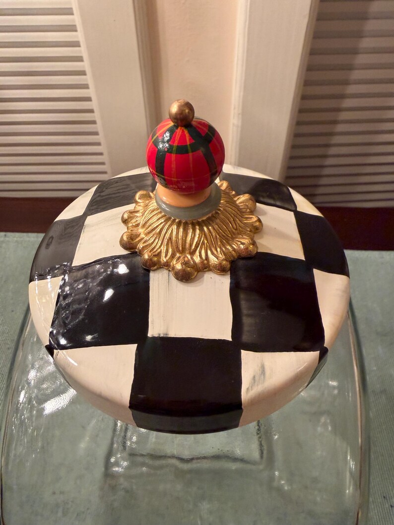 May include: A glass jar lid featuring a black and white checkered pattern. The lid is topped with a gold-colored decorative base and a small, spherical finial painted with a red, green, and black plaid design.