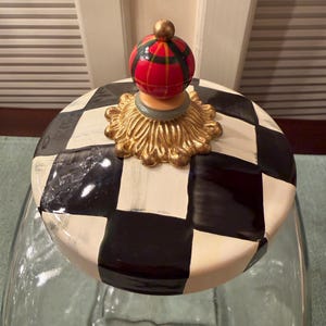 May include: A glass jar lid featuring a black and white checkered pattern. The lid is topped with a decorative gold-colored base and a spherical finial painted with a red, green, and yellow plaid design, finished with a small gold-colored bead.