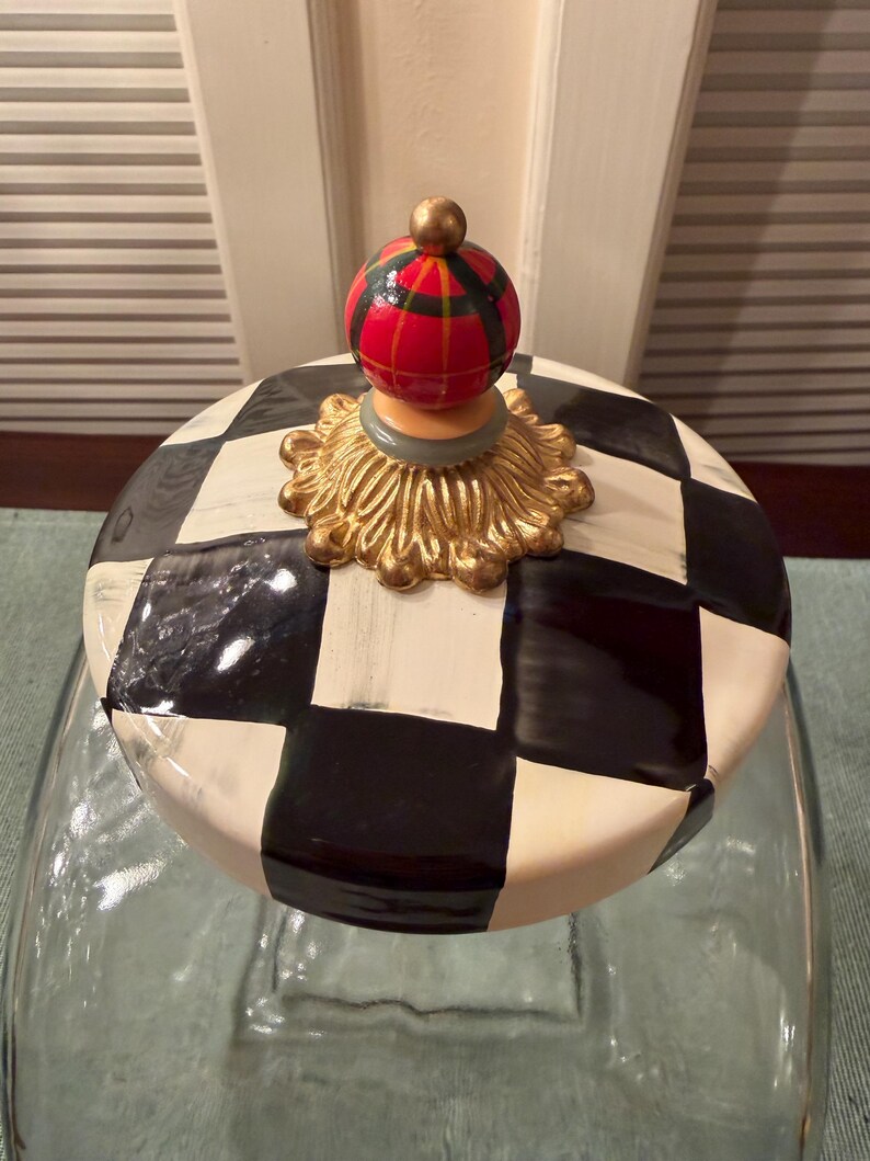May include: A glass jar lid featuring a black and white checkered pattern. The lid is topped with a decorative gold-colored base and a spherical finial painted with a red, green, and yellow plaid design, finished with a small gold-colored bead.