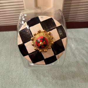 May include: A clear glass storage jar with a round lid featuring a black and white checkered pattern. The center of the lid is adorned with a gold-toned, sunburst-shaped decorative knob that has a small, red and green plaid circular accent.