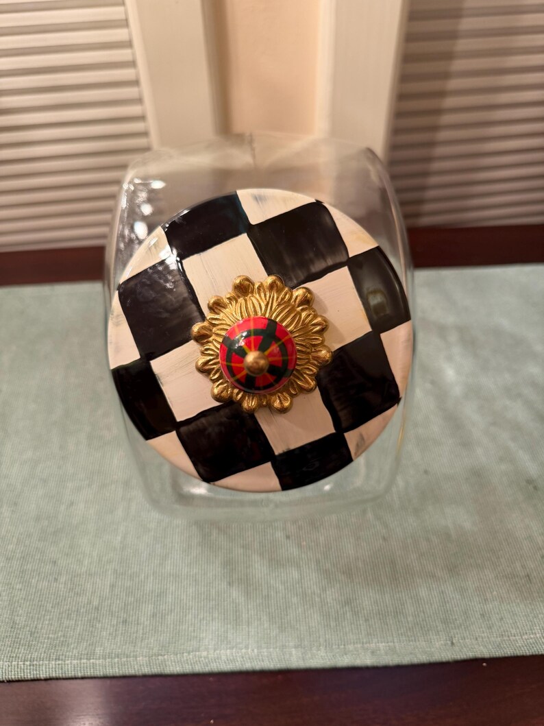 May include: A clear glass storage jar with a round lid featuring a black and white checkered pattern. The center of the lid is adorned with a gold-toned, sunburst-shaped decorative knob that has a small, red and green plaid circular accent.