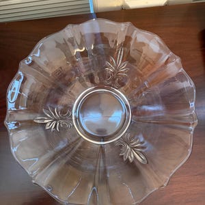 May include: A clear glass serving bowl with a scalloped edge and a central, circular design. The bowl features decorative leaf-like etchings around the center. The bowl is sitting on a dark brown surface.