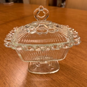May include: A clear glass candy dish with a matching lid, featuring a rectangular shape with vertical ribbing, a scalloped beaded edge, and a central loop handle on top. The dish is supported by a rectangular pedestal base made of clear glass.