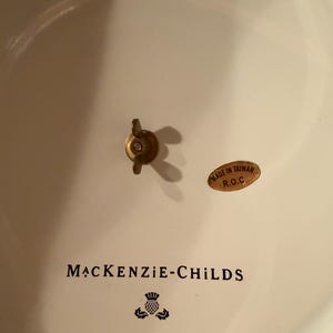 May include: The underside of a cream-colored ceramic piece featuring the black text MACKENZIE-CHILDS, a thistle logo, and the number 6997TW13243. A brass-colored wing nut is centered, and an oval sticker reads MADE IN TAIWAN R.O.C.