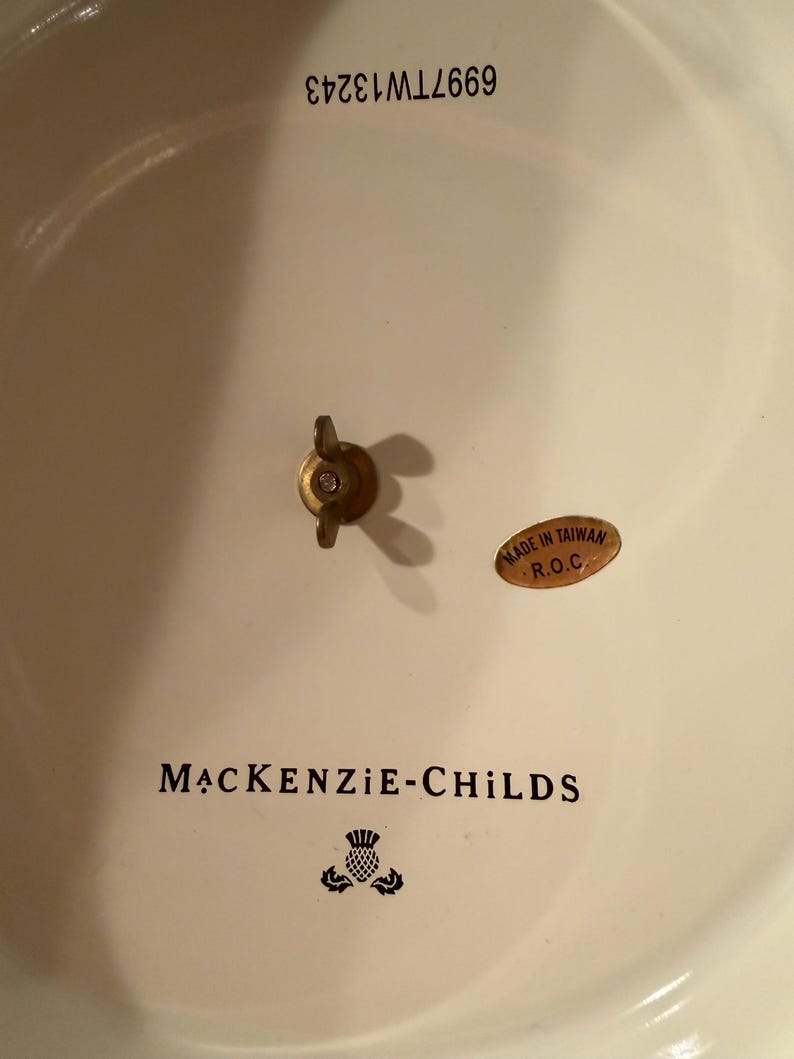 May include: The underside of a cream-colored ceramic piece featuring the black text MACKENZIE-CHILDS, a thistle logo, and the number 6997TW13243. A brass-colored wing nut is centered, and an oval sticker reads MADE IN TAIWAN R.O.C.