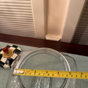 May include: A clear glass jar with a square opening, shown with a yellow measuring tape across the top indicating a diameter of approximately 6 inches. Beside the jar sits a lid featuring a black and white checkered pattern and a red finial.