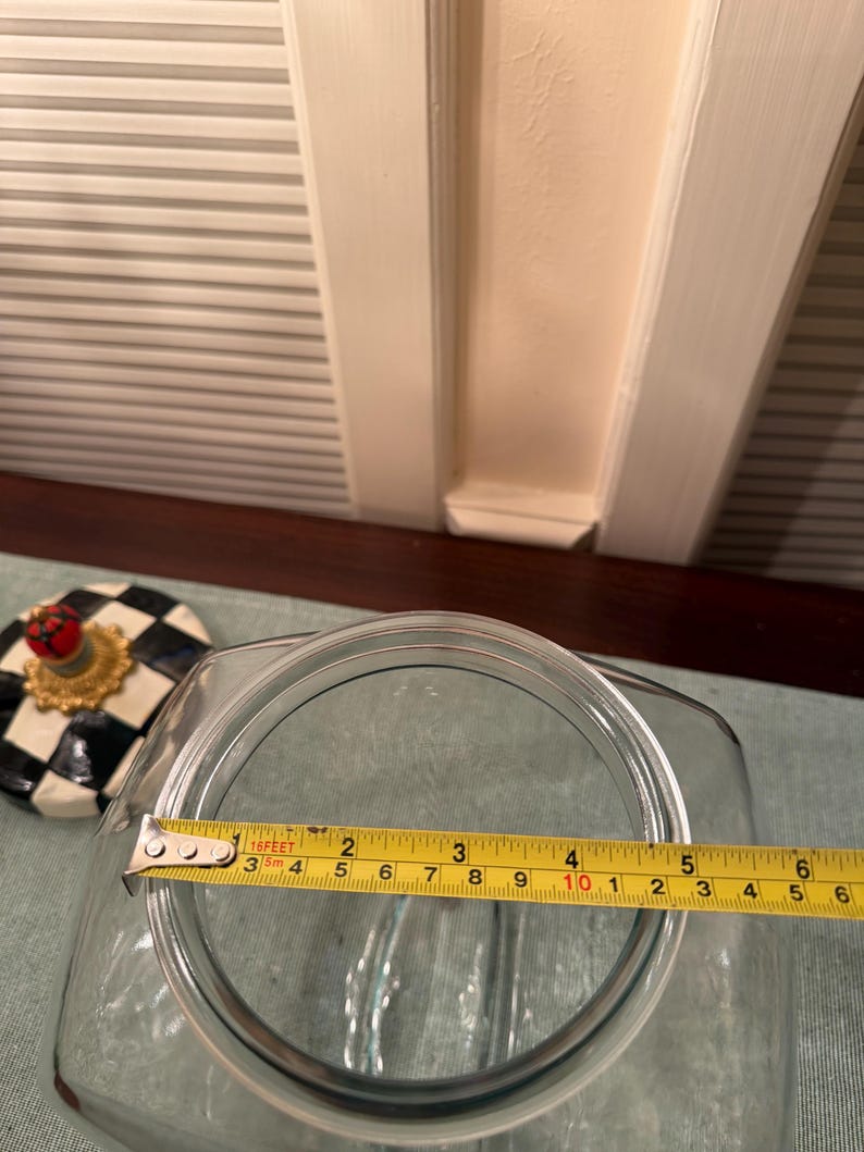 May include: A clear glass jar with a square opening, shown with a yellow measuring tape across the top indicating a diameter of approximately 6 inches. Beside the jar sits a lid featuring a black and white checkered pattern and a red finial.