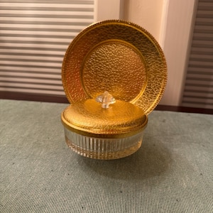 May include: A round glass container with a clear, vertically ribbed base and a textured gold lid featuring a small, clear, pointed knob handle. Behind the container stands a matching round, textured gold plate or tray with a decorative edge.