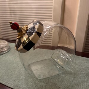 May include: A clear glass canister with a round body and a decorative lid. The lid features a black and white checkered pattern, a gold-toned ornate base, and a small red and black plaid sphere finial at the very top of the lid structure.