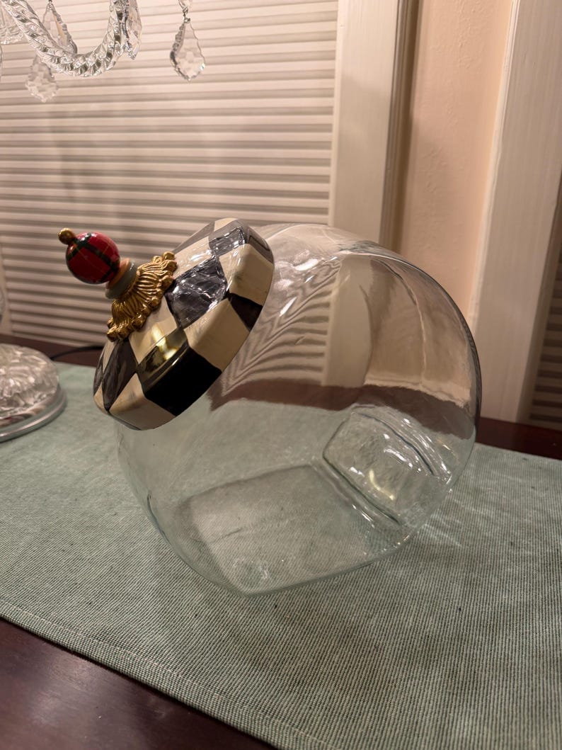 May include: A clear glass canister with a round body and a decorative lid. The lid features a black and white checkered pattern, a gold-toned ornate base, and a small red and black plaid sphere finial at the very top of the lid structure.
