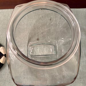 May include: A clear glass storage jar with a wide, circular opening and a square-shaped base. The jar features a thick, reinforced rim designed for a lid, with smooth, transparent walls that allow for a clear view into the interior space.