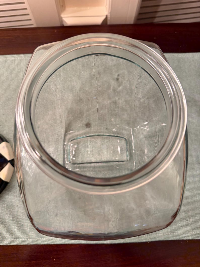 May include: A clear glass storage jar with a wide, circular opening and a square-shaped base. The jar features a thick, reinforced rim designed for a lid, with smooth, transparent walls that allow for a clear view into the interior space.