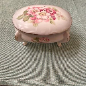 May include: A small, round, white ceramic trinket box with a lid. The lid is decorated with a floral design featuring pink and white flowers and green leaves. The box has three small feet and a gold trim.