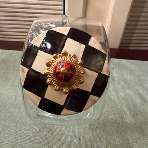 May include: A clear glass canister with a round lid featuring a black and white checkerboard pattern. The lid is topped with a gold-toned, scalloped metal base and a small, round, multi-colored knob with red, green, and black segments.