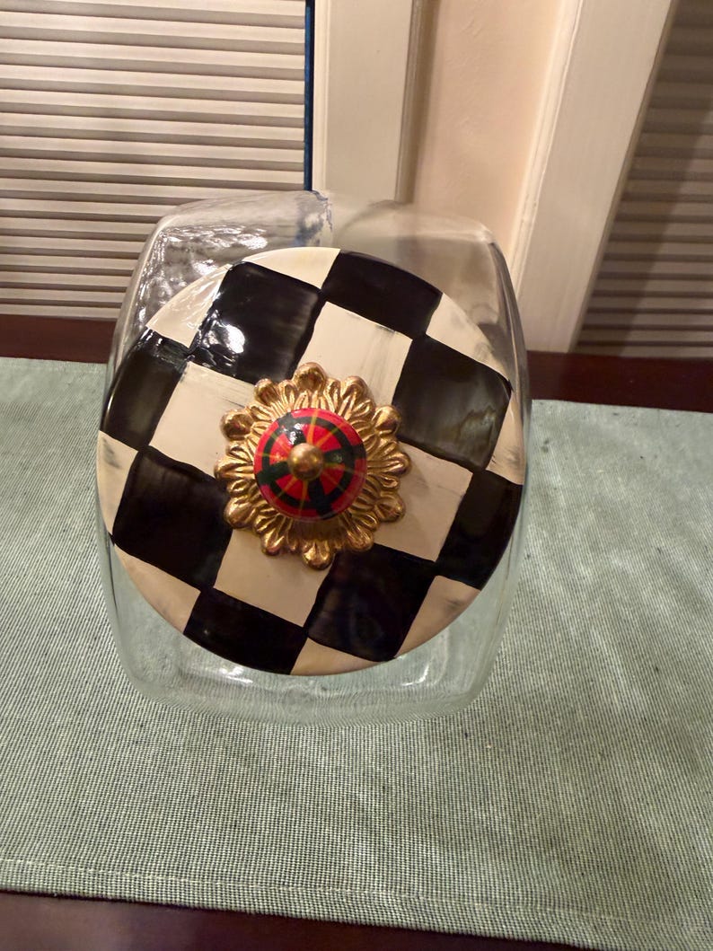 May include: A clear glass canister with a round lid featuring a black and white checkerboard pattern. The lid is topped with a gold-toned, scalloped metal base and a small, round, multi-colored knob with red, green, and black segments.