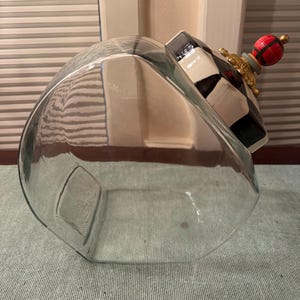 May include: A clear glass canister with a slanted opening and a decorative lid. The lid features a black and white checkered pattern, a gold-toned base, and a round finial painted with a red, green, and black plaid design and a gold tip.