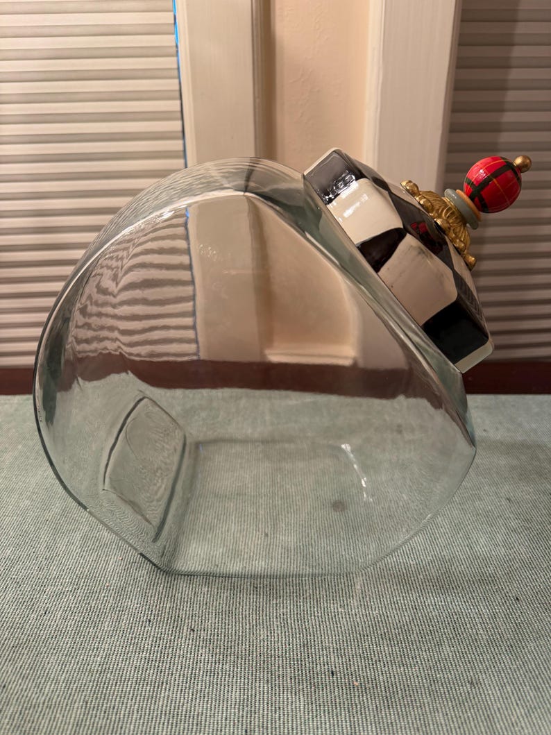 May include: A clear glass canister with a slanted opening and a decorative lid. The lid features a black and white checkered pattern, a gold-toned base, and a round finial painted with a red, green, and black plaid design and a gold tip.