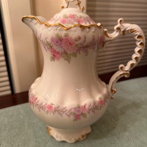 May include: A white porcelain pitcher with a lid, adorned with pink floral patterns and gold trim. The handle is ornate, and the pitcher has a scalloped base. The overall design is vintage and elegant, suitable for serving beverages.