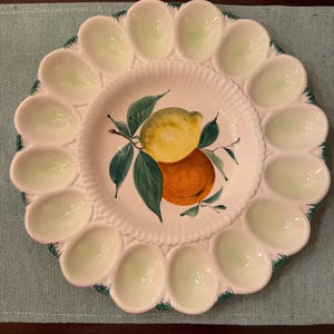 May include: White ceramic egg serving platter with 12 egg-shaped indentations around the rim. The center features a painted design of a lemon and orange with green leaves. The rim has a green trim.
