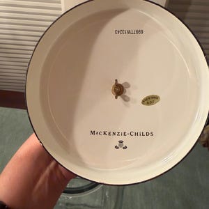 May include: The underside of a round, cream-colored MacKenzie-Childs lid featuring a central brass wing nut, a gold sticker reading Made in Taiwan R.O.C., and the text 6997TW13243. The brand name MacKenzie-Childs is printed in black with a thistle logo.