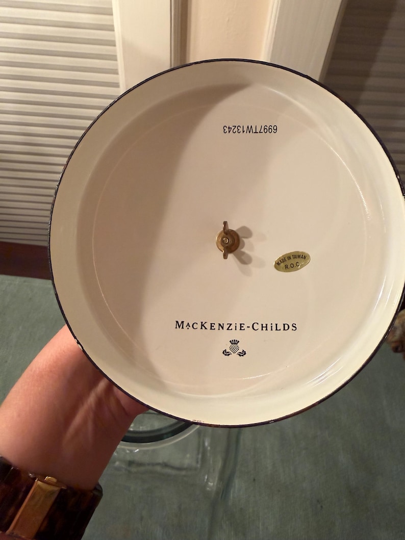 May include: The underside of a round, cream-colored MacKenzie-Childs lid featuring a central brass wing nut, a gold sticker reading Made in Taiwan R.O.C., and the text 6997TW13243. The brand name MacKenzie-Childs is printed in black with a thistle logo.