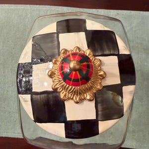 May include: A clear glass canister with a round lid featuring a black and white checkered pattern. The lid is topped with a decorative gold-toned metal base and a small, rounded knob painted with a red, green, and black plaid-like design.