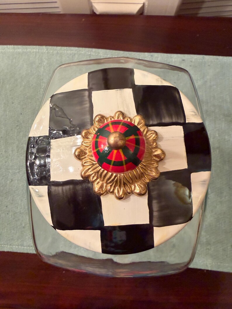 May include: A clear glass canister with a round lid featuring a black and white checkered pattern. The lid is topped with a decorative gold-toned metal base and a small, rounded knob painted with a red, green, and black plaid-like design.