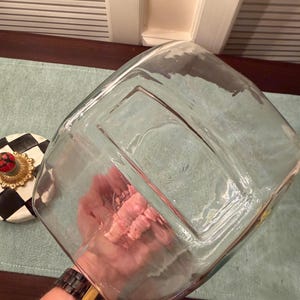 May include: A clear, square glass container with a thick base and rounded corners. Beside it sits a circular lid featuring a black and white checkered pattern, a gold-toned decorative rim, and a small red and green knob at the center.