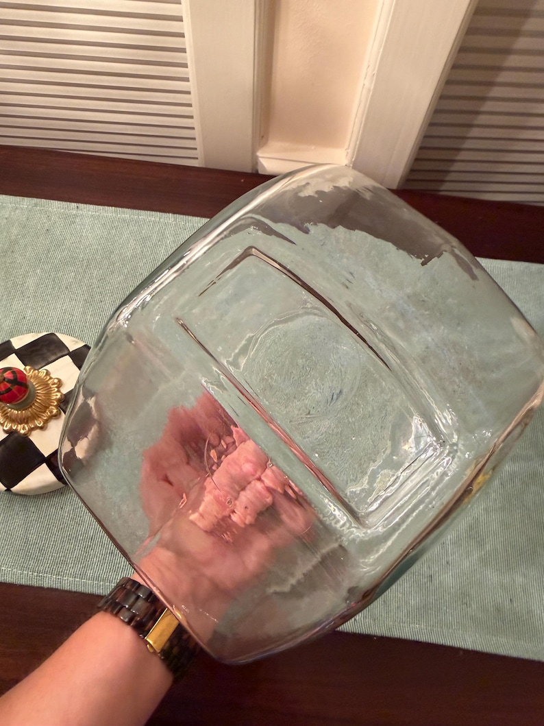 May include: A clear, square glass container with a thick base and rounded corners. Beside it sits a circular lid featuring a black and white checkered pattern, a gold-toned decorative rim, and a small red and green knob at the center.