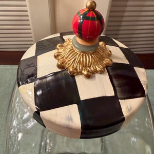 May include: A clear glass jar with a lid featuring a black and white checkered pattern. The lid is topped with a gold-colored decorative base and a small, round finial painted with red, green, and black plaid stripes and a gold-colored tip.