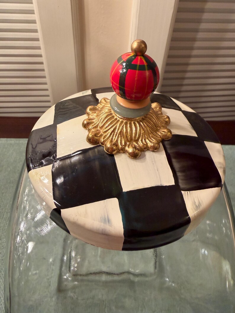 May include: A clear glass jar with a lid featuring a black and white checkered pattern. The lid is topped with a gold-colored decorative base and a small, round finial painted with red, green, and black plaid stripes and a gold-colored tip.