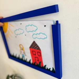 Könnte beinhalten: Ein blauer Bilderrahmen mit einer Kinderzeichnung eines Hauses, einer Person, einer Sonne und Wolken. Die Zeichnung ist mit Buntstiften und Markern erstellt. Der Rahmen ist tiefblau und an einer weißen Wand befestigt.
