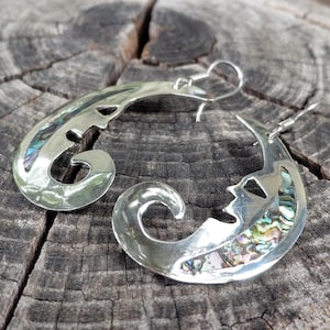 May include: A pair of crescent moon-shaped earrings crafted from polished silver-tone metal. Each earring features a stylized face profile and an inlay of iridescent abalone shell with swirling green, blue, and purple hues, finished with silver-tone ear wires.