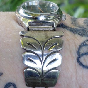 May include: A silver-toned wristwatch with a black face and orange hands. The watch band features a stylized leaf design. The watch is a vintage accessory.