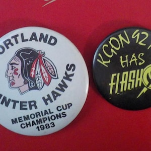 May include: Two vintage pinback buttons. One is white with "Portland Winter Hawks" and a Native American head logo. The other is black with "KCON 92 FM HAS FLASH" in yellow. Both are round and likely from the 1980s.