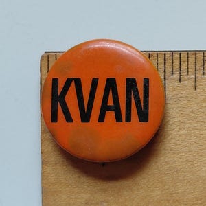 May include: A round, orange button with the black letters "KVAN" printed on it. The button is next to a wooden ruler.