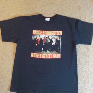 May include: Black t-shirt with a photo of Bruce Springsteen and The E Street Band. The text "Bruce Springsteen & The E Street Band" is printed in orange and red on the shirt.