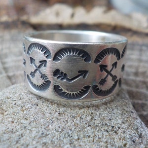 May include: A silver band ring with a repeating pattern of crossed arrows and crescent shapes.