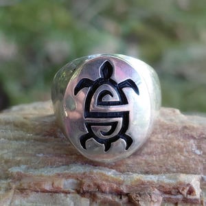 May include: A polished silver ring featuring a circular face with a recessed, black-filled turtle motif. The turtle design is stylized with geometric, swirling patterns within its shell and limbs, centered on the smooth, rounded metal band.