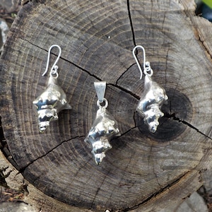 May include: A set of silver earrings and a pendant featuring a detailed seashell design. The jewelry is displayed on a wooden surface.