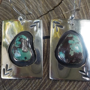 May include: A pair of silver earrings with turquoise stones set in a rectangular frame. The earrings have a simple design with a small arrow motif on each side.