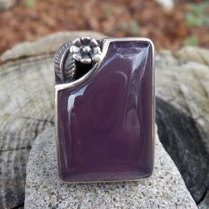 May include: A silver ring with a rectangular purple stone and a floral design on the side.