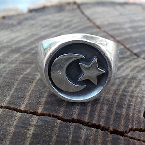 May include: A silver ring featuring a crescent moon and a star design. The ring has a rounded face with the symbols set against a dark background. The ring is displayed on a textured wooden surface.