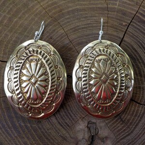 May include: A pair of gold oval-shaped earrings with a detailed floral design. The earrings have a silver hook closure.