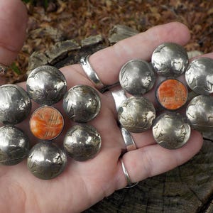 May include: A silver and orange flower-shaped brooch made from repurposed US Indian Head nickels. The center of the flower is a large orange stone.