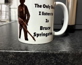May include: White ceramic mug with a black silhouette of a person holding a guitar. The mug features the text "The Only Boss I listen to Is Bruce Springsteen". The mug has a curved handle.